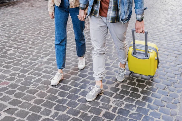 Обрезанный вид пары, держащейся за руки и путешествующей с чемоданом вместе — стоковое фото