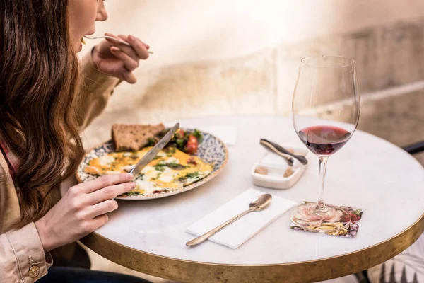 Vista parcial de la mujer comiendo tortilla con copa de vino tinto en la cafetería - foto de stock