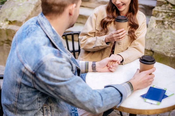 Cropped view of couple with paper cups of coffee holding hands in cafe in city — Stock Photo