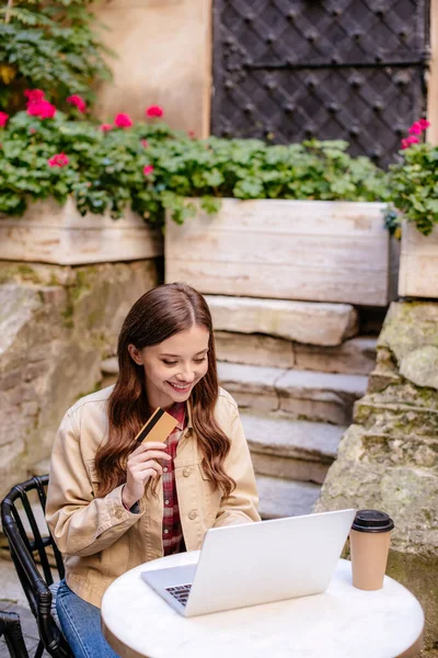 Selective focus of woman smiling, holding credit card and using laptop in cafe near stone building in city — Stock Photo