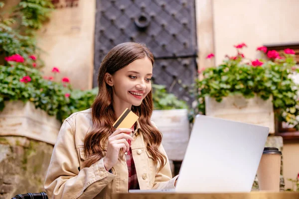 Foco seletivo da mulher sorrindo, segurando cartão de crédito e usando laptop no café na cidade — Fotografia de Stock