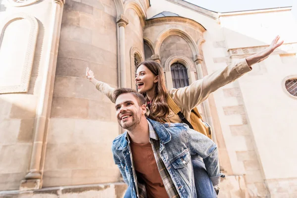 Low angle view of man piggybacking girlfriend with open arms near building in city — Stock Photo