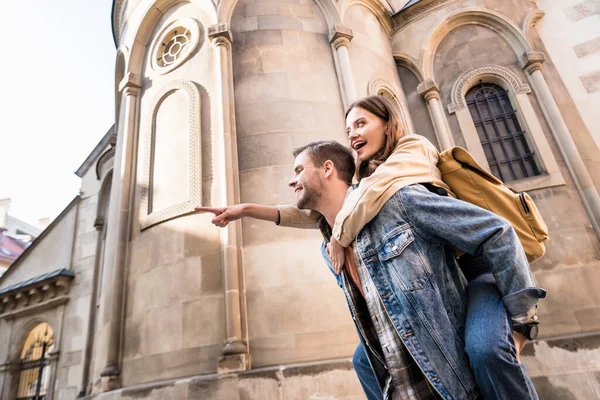 Low angle view of man piggybacking girlfriend pointing with finger near building in city — Stock Photo