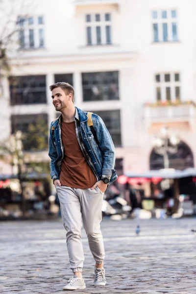 Homme avec sac à dos et les mains dans les poches souriant et marchant dans la ville — Photo de stock