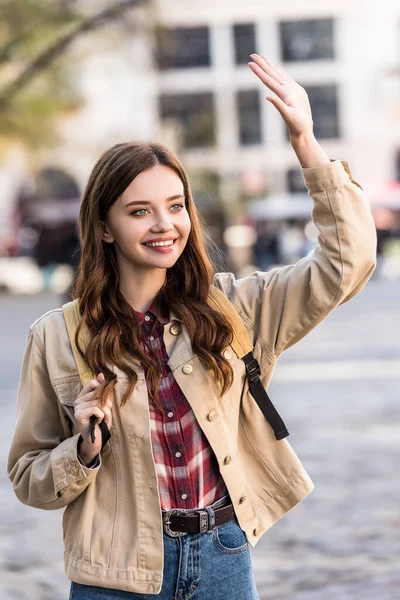 Hermosa mujer saludando de la mano y sonriendo con la mochila en la ciudad - foto de stock