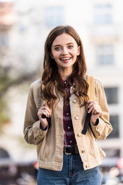 Menina atraente olhando para a câmera e sorrindo com mochila na cidade — Fotografia de Stock