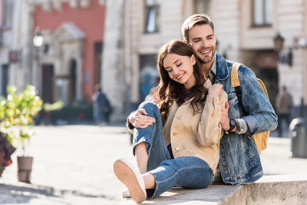 Enfoque selectivo de novio abrazando chica sentada en la superficie de piedra y sonriendo en la ciudad - foto de stock