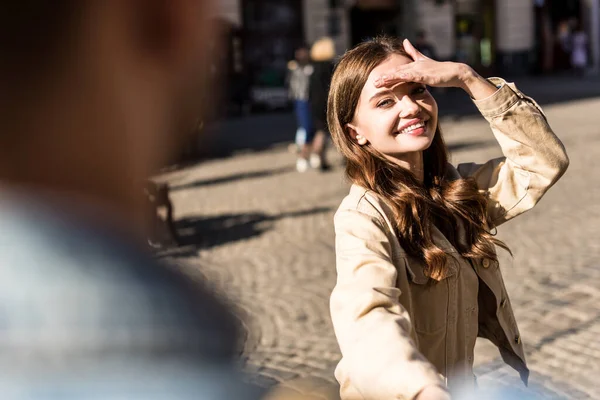 Cropped view of girl smiling, covering face from sunlight and holding male hand in city — Stock Photo