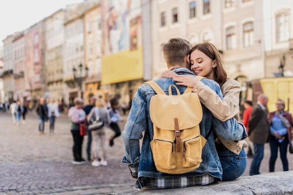 Freund mit Rucksack und Freundin mit geschlossenen Augen umarmen und lächeln in der Stadt — Stockfoto