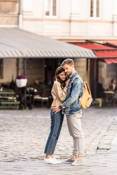 Pareja feliz abrazando y sonriendo con mochila en la ciudad en Europa - foto de stock