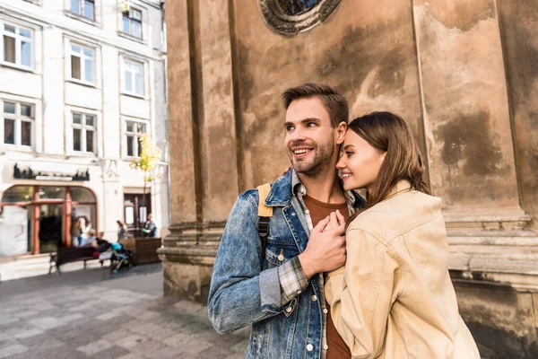Foco seletivo de namorada e namorado olhando para longe, de mãos dadas e sorrindo perto da parede na cidade — Fotografia de Stock