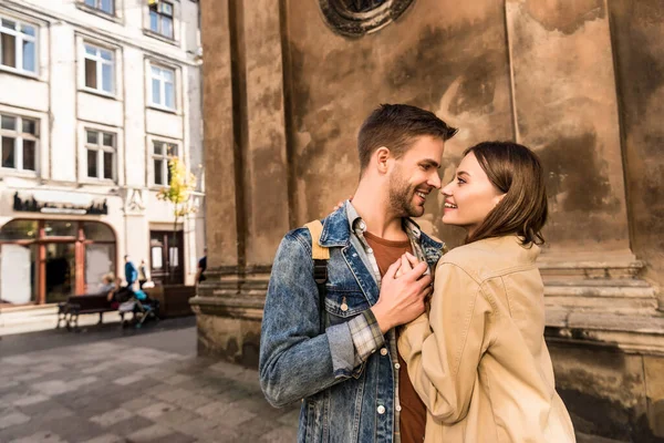 Hombre y mujer tomados de la mano, mirándose el uno al otro y sonriendo cerca de la pared en la ciudad - foto de stock