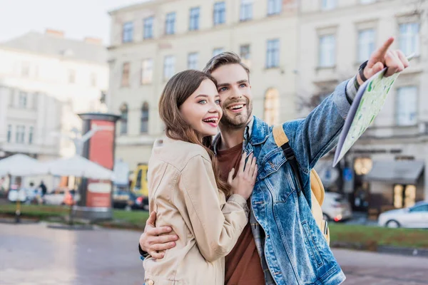 Man smiling, holding map, pointing with finger and hugging happy woman in city — Stock Photo