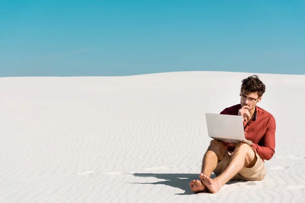 Pensive handsome freelancer on sandy beach with laptop against clear blue sky — Stock Photo