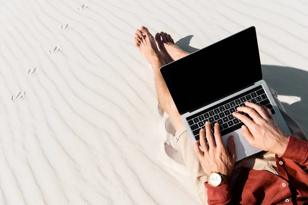 Vista recortada del freelancer sentado en la playa de arena con el ordenador portátil contra el cielo azul claro - foto de stock