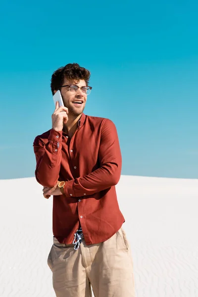 Счастливый красавец на песчаном пляже, разговаривающий по смартфону на фоне ясного голубого неба — стоковое фото