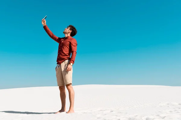 Bell'uomo sulla spiaggia sabbiosa che parla sullo smartphone contro il cielo blu chiaro — Foto stock