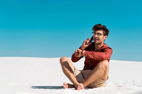 Счастливый человек на песчаном пляже разговаривает по смартфону на фоне ясного голубого неба — стоковое фото