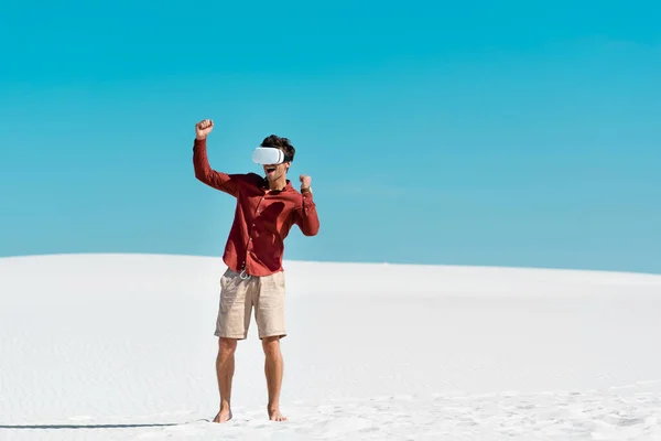 Excited handsome man on sandy beach in vr headset against clear blue sky — Stock Photo