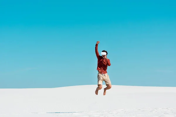 Man on sandy beach in vr headset jumping against clear blue sky — Stock Photo