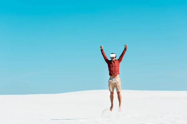 Uomo sulla spiaggia sabbiosa in auricolare vr saltando contro il cielo blu chiaro — Foto stock