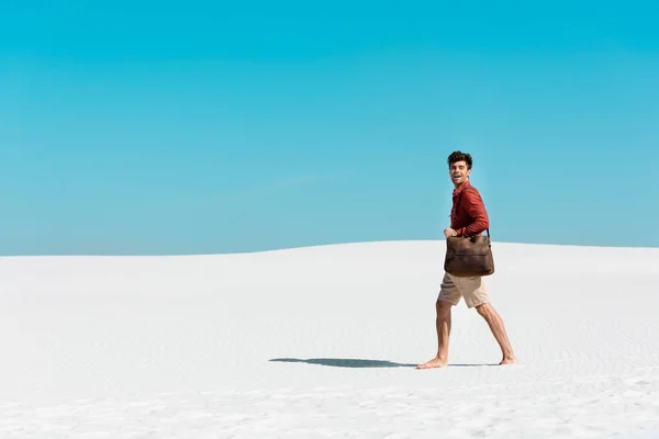 Vista laterale di bell'uomo con borsa in pelle che cammina sulla spiaggia sabbiosa contro il cielo blu chiaro — Foto stock