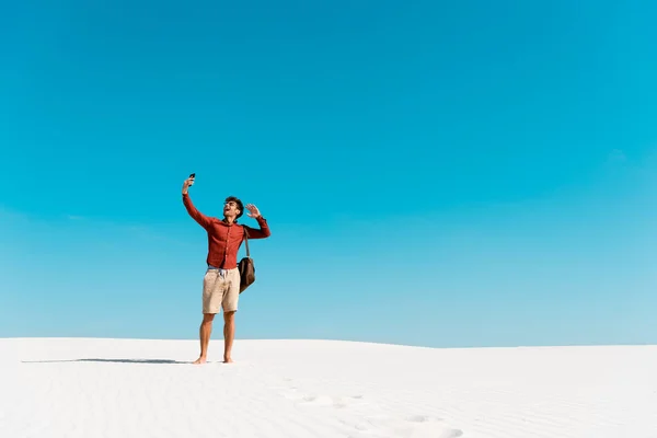 Hombre guapo con bolsa de cuero usando teléfono inteligente en la playa de arena contra el cielo azul claro — Stock Photo