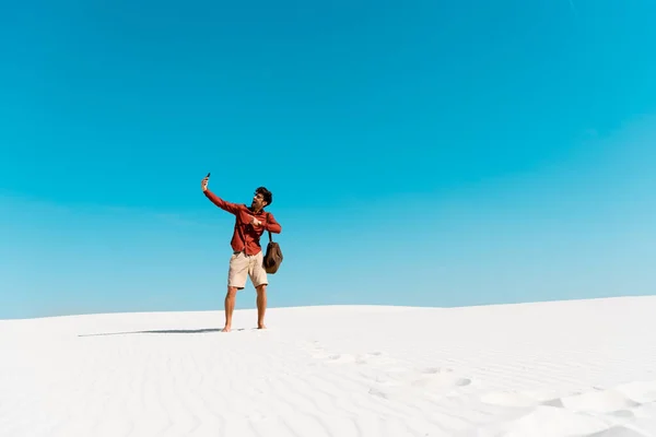 Красивий чоловік зі шкіряною сумкою бере селфі на смартфон на піщаному пляжі проти прозорого блакитного неба — стокове фото