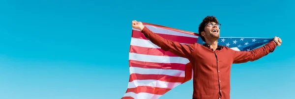 Vista de ángulo bajo del hombre feliz con bandera americana contra el viento y el cielo azul claro, tiro panorámico - foto de stock