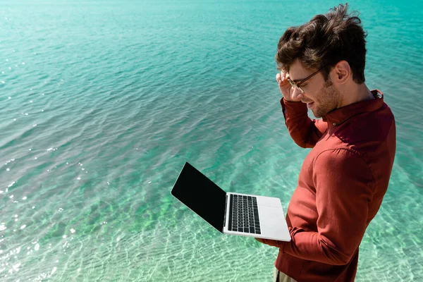 Side view of smiling handsome freelancer with laptop standing in turquoise water — Stock Photo