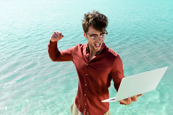 Excitado guapo freelancer con portátil de pie en agua turquesa - foto de stock