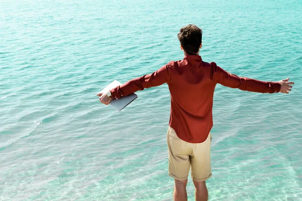 Back view of freelancer with laptop in outstretched hands standing in turquoise water — Stock Photo