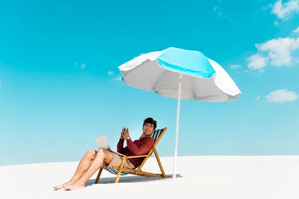 Smiling freelancer sitting with laptop in deck chair under umbrella on sandy beach against blue sky — Stock Photo