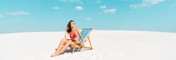 Sourire belle fille sexy en maillot de bain et lunettes de soleil assis dans la chaise longue sur la plage de sable avec ciel bleu et nuages, vue panoramique — Photo de stock