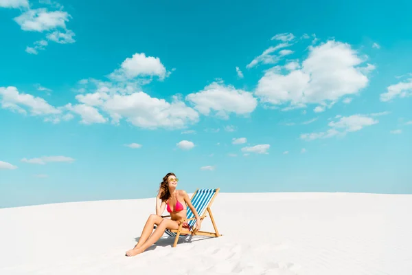 Smiling beautiful sexy girl in swimsuit and sunglasses sitting in deck chair on sandy beach with blue sky and clouds — Stock Photo