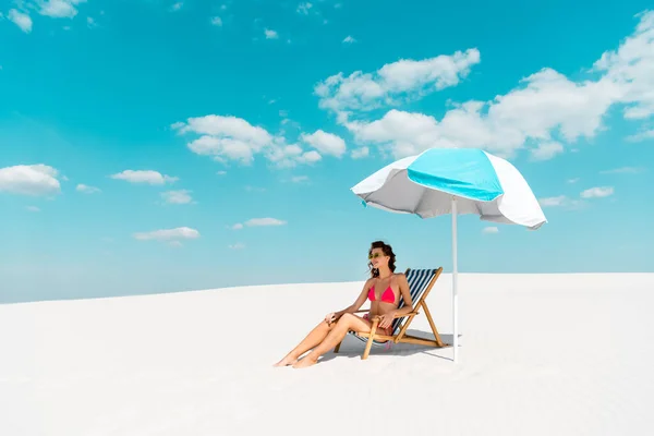 Улыбающаяся красивая сексуальная девушка в купальнике и солнцезащитных очках, сидящая в шезлонге под зонтиком на песчаном пляже — стоковое фото