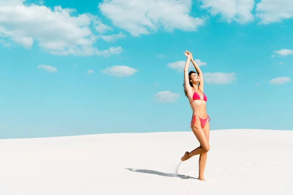 Beautiful sexy girl in swimsuit with hands in air on sandy beach with blue sky and clouds — Stock Photo