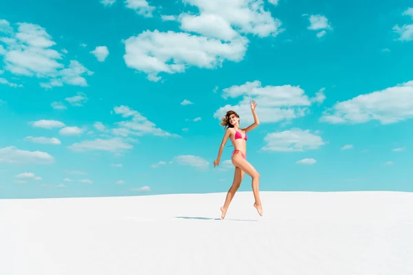 Улыбающаяся красивая сексуальная девушка в купальнике, прыгающая по песчаному пляжу с голубым небом и облаками — стоковое фото