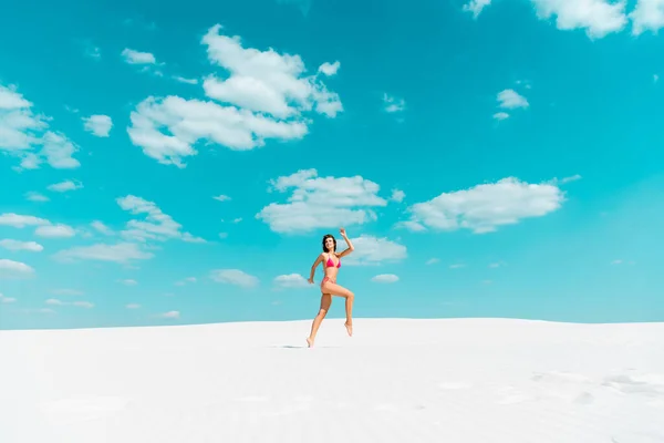 Beautiful sexy girl in swimsuit jumping on sandy beach with blue sky and clouds — Stock Photo