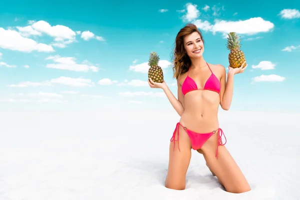 Smiling beautiful sexy girl in swimsuit with pineapples on sandy beach with blue sky and clouds — Stock Photo