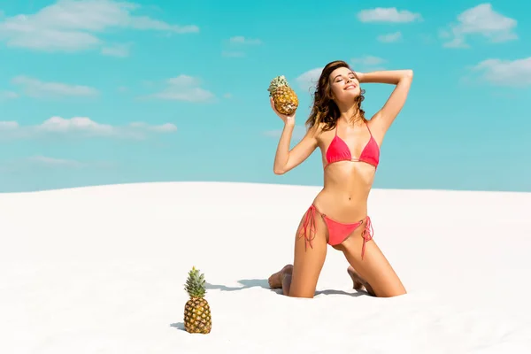 Smiling beautiful sexy girl in swimsuit with pineapples on sandy beach with blue sky and clouds — Stock Photo