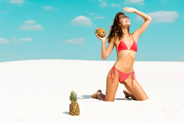 Smiling beautiful sexy girl in swimsuit with pineapples hiding from sun on sandy beach with blue sky and clouds — Stock Photo