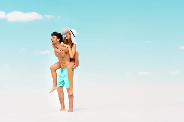 Sonriente joven hombre piggybacking novia en la playa de arena - foto de stock