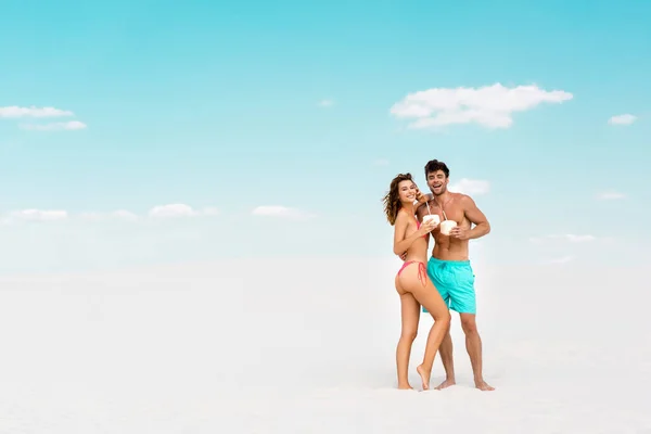 Sonriente joven pareja abrazando mientras sostiene bebidas de coco en la playa de arena - foto de stock