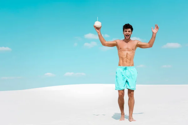 Homem sexy feliz com torso muscular em shorts de natação com bebida de coco na praia de areia — Fotografia de Stock
