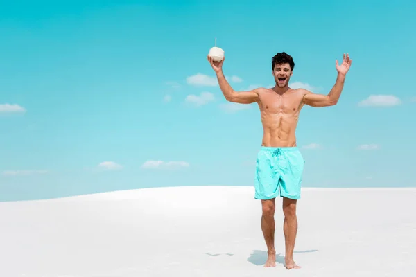 Happy sexy man with muscular torso in swim shorts with coconut drink on sandy beach — Stock Photo