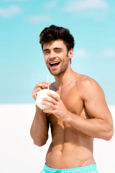 Glücklich sexy Mann mit muskulösem Oberkörper in Badehosen mit Kokosgetränk am Sandstrand — Stockfoto