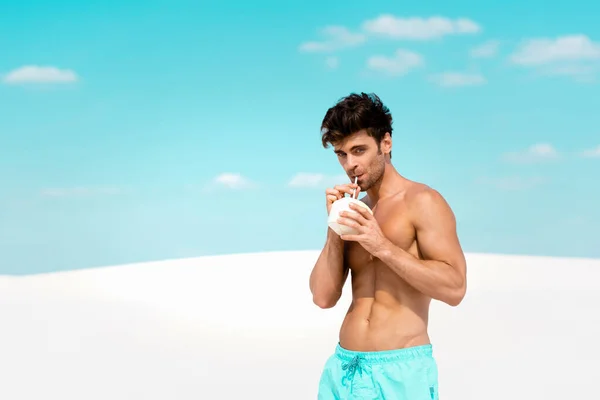 Sexy man with muscular torso in swim shorts with coconut drink on sandy beach — Stock Photo