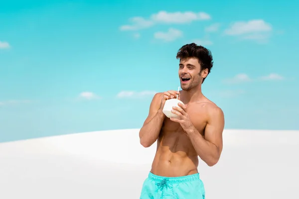 Uomo sexy sorridente con busto muscoloso in pantaloncini da bagno con bevanda al cocco sulla spiaggia sabbiosa — Foto stock