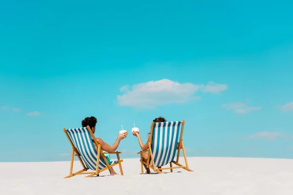 Vista trasera de pareja joven sentada en tumbonas con bebidas de coco en la playa de arena - foto de stock